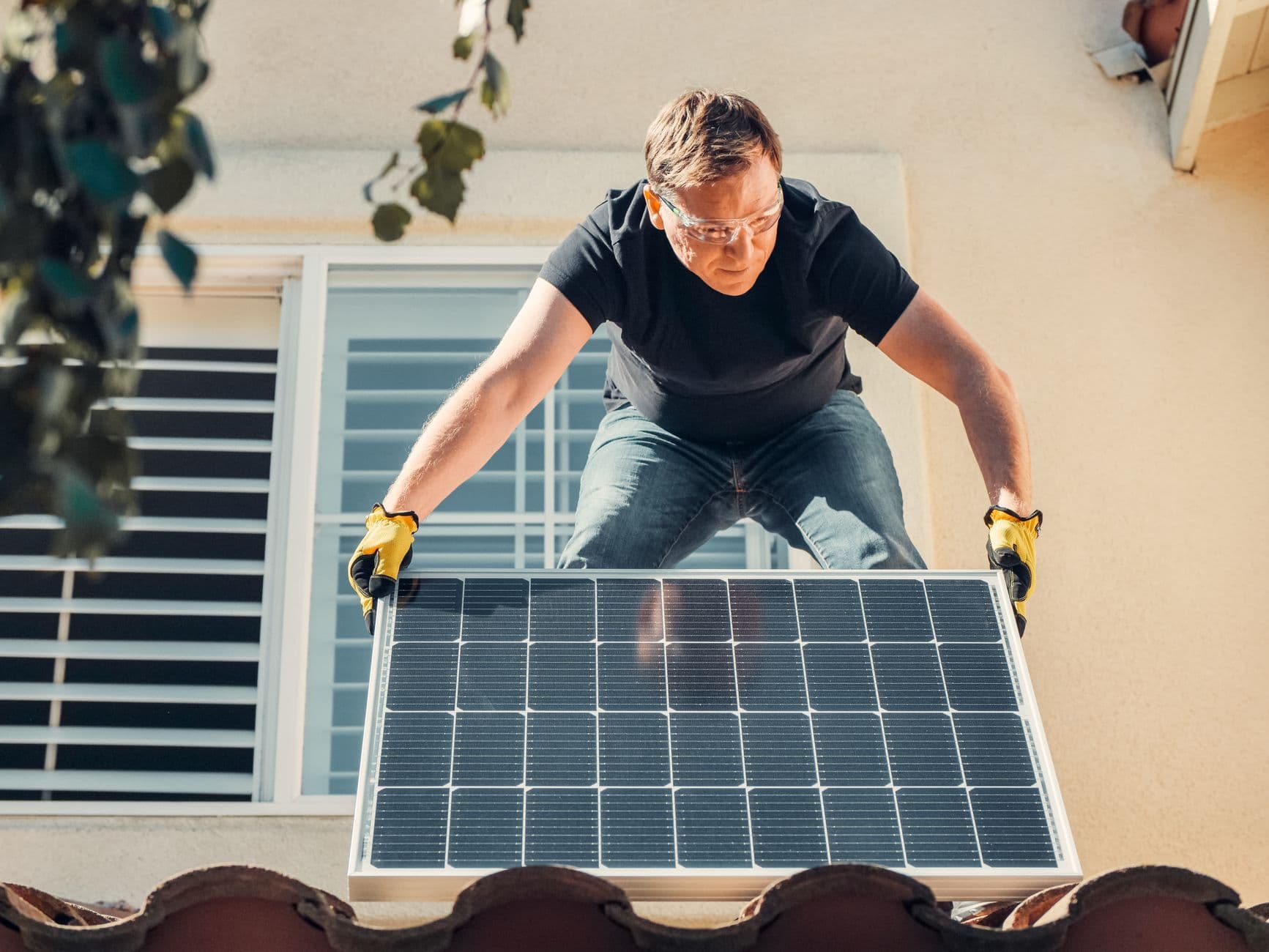An installer in safety glasses and gloves lifting a solar panel onto a tiled roof